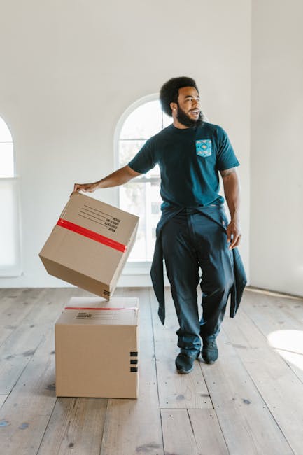 Two movers from Removal Van Brent are standing outside a storefront with Asian signage, preparing to load or unload items from a large, partially open delivery truck. One mover is holding a collapsibl